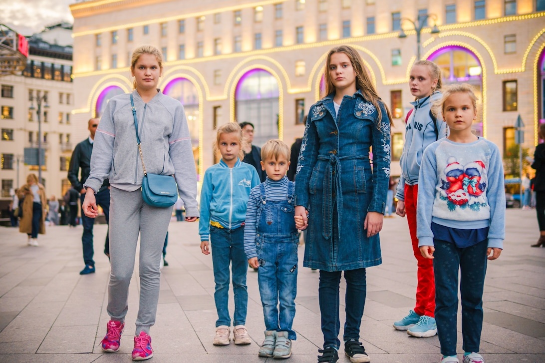 Eine Gruppe von Kindern und Teenagern in modischer Freizeitkleidung geht durch eine städtische Umgebung mit beleuchteten Gebäuden.
